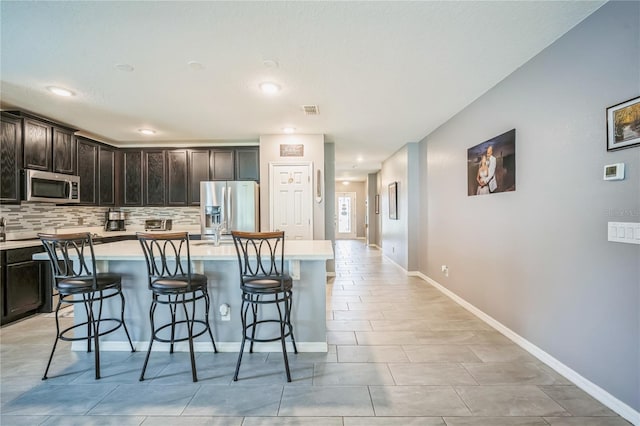 kitchen featuring a breakfast bar area, an island with sink, backsplash, appliances with stainless steel finishes, and dark brown cabinetry