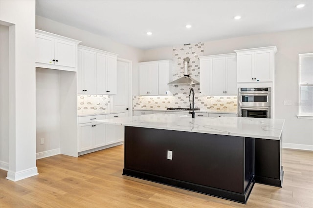 kitchen with light wood-type flooring, an island with sink, wall chimney exhaust hood, appliances with stainless steel finishes, and white cabinets