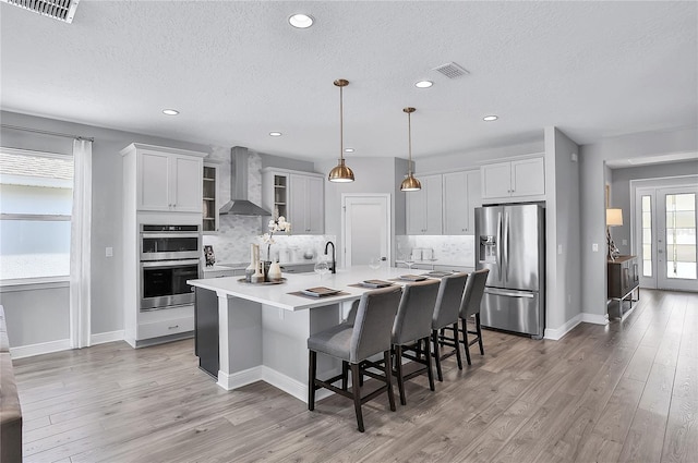 kitchen featuring light wood-type flooring, appliances with stainless steel finishes, wall chimney range hood, an island with sink, and a kitchen bar