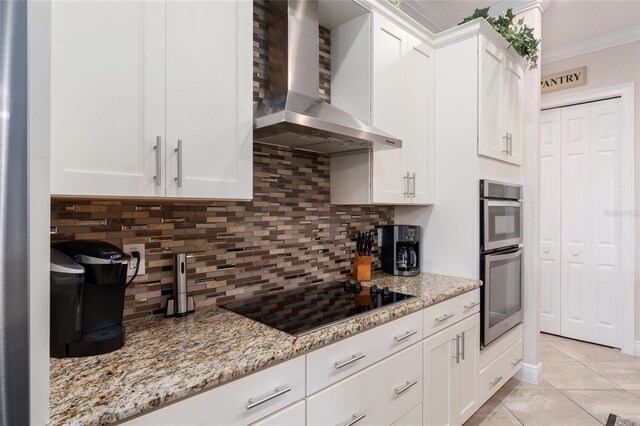 kitchen with black electric stovetop, ornamental molding, light stone counters, white cabinets, and wall chimney range hood