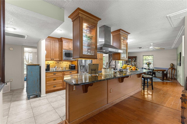 kitchen featuring a breakfast bar area, light wood-type flooring, a textured ceiling, ceiling fan, and wall chimney range hood