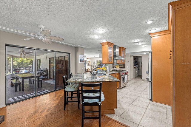 kitchen with ceiling fan, a textured ceiling, wall chimney range hood, appliances with stainless steel finishes, and light wood-type flooring