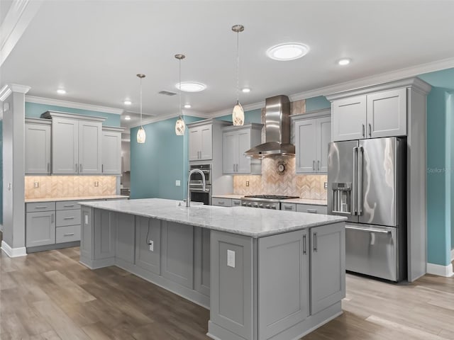 kitchen featuring appliances with stainless steel finishes, hanging light fixtures, light stone counters, wall chimney exhaust hood, and a kitchen island with sink