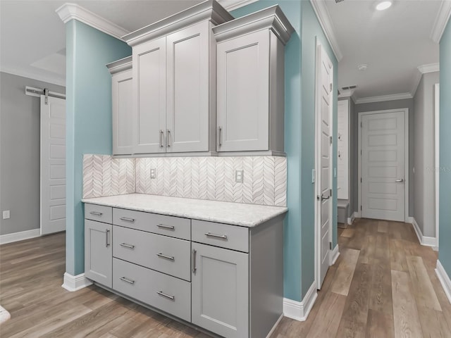 kitchen featuring light wood-type flooring, a barn door, decorative backsplash, crown molding, and light stone countertops