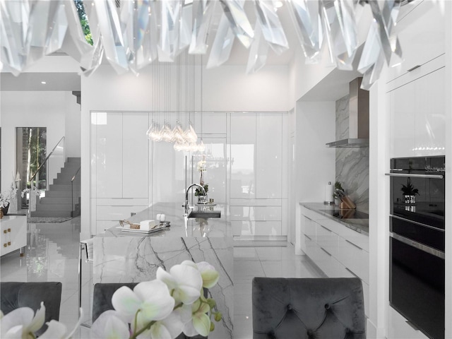 kitchen with black electric cooktop, white cabinetry, light stone counters, sink, and wall chimney range hood