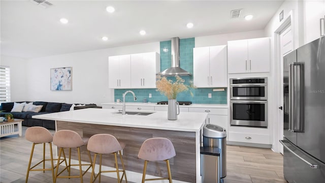 kitchen with a kitchen island with sink, sink, wall chimney range hood, light hardwood / wood-style flooring, and stainless steel appliances