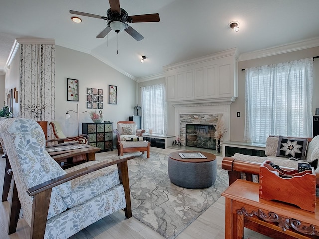 living room featuring ceiling fan, light wood-type flooring, vaulted ceiling, a stone fireplace, and crown molding