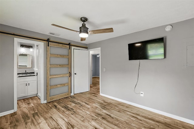 unfurnished bedroom featuring ceiling fan, sink, a barn door, ensuite bathroom, and light wood-type flooring