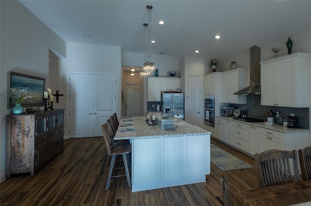 kitchen with a kitchen island with sink, wall chimney range hood, pendant lighting, white cabinetry, and appliances with stainless steel finishes