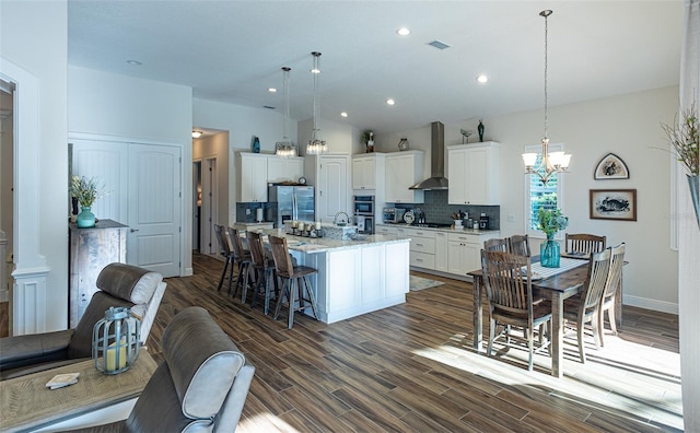 kitchen featuring wall chimney range hood, dark hardwood / wood-style floors, an island with sink, hanging light fixtures, and white cabinetry