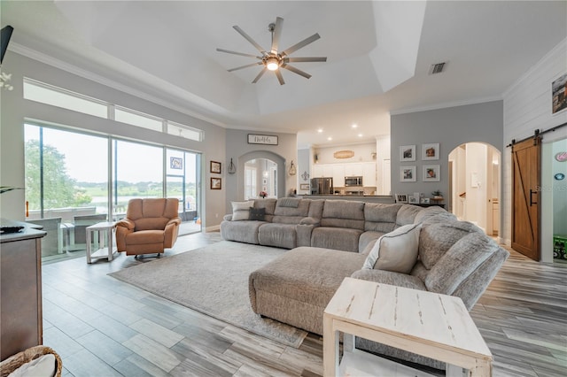 living room with ceiling fan, ornamental molding, a tray ceiling, a barn door, and light hardwood / wood-style floors