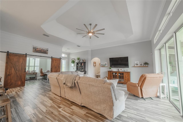 living room with light hardwood / wood-style floors, a barn door, crown molding, a tray ceiling, and ceiling fan