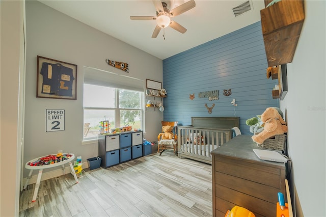 bedroom with light wood-type flooring, wooden walls, ceiling fan, and a nursery area