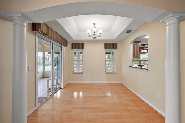 unfurnished dining area with light hardwood / wood-style flooring, ornate columns, ceiling fan with notable chandelier, and a raised ceiling