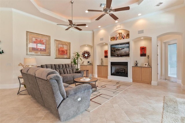 tiled living room with crown molding, a raised ceiling, built in features, and ceiling fan