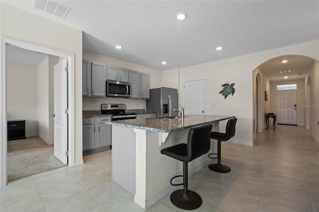 kitchen with gray cabinetry, stainless steel appliances, dark stone countertops, and a center island with sink