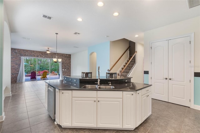 kitchen featuring stainless steel dishwasher, hanging light fixtures, white cabinetry, a kitchen island with sink, and brick wall