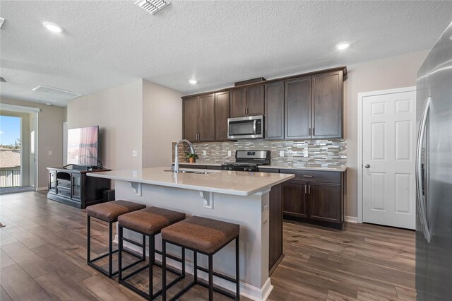 kitchen with dark brown cabinetry, a center island with sink, sink, stainless steel appliances, and dark hardwood / wood-style floors