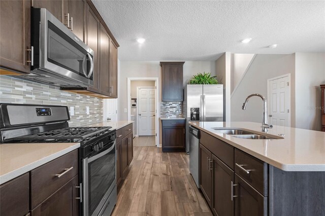 kitchen with light wood-type flooring, a kitchen island with sink, sink, appliances with stainless steel finishes, and backsplash