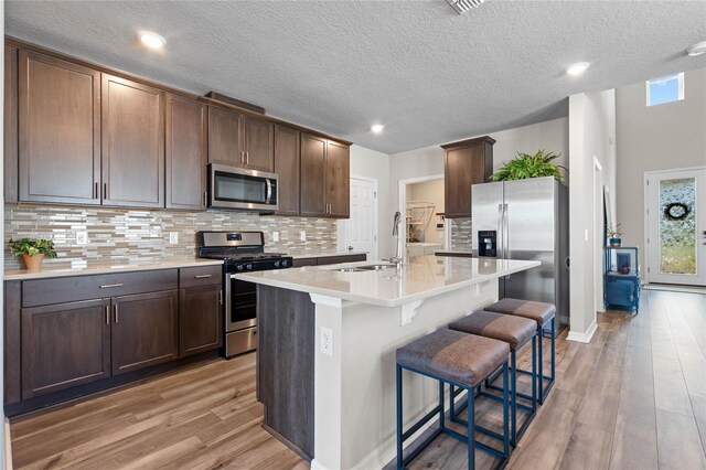 kitchen with an island with sink, light wood-type flooring, dark brown cabinetry, appliances with stainless steel finishes, and a textured ceiling