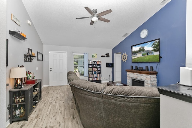 living room featuring ceiling fan, lofted ceiling, a stone fireplace, a textured ceiling, and light hardwood / wood-style flooring