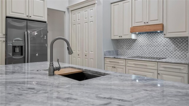 kitchen featuring stainless steel fridge, sink, backsplash, black electric stovetop, and light stone countertops