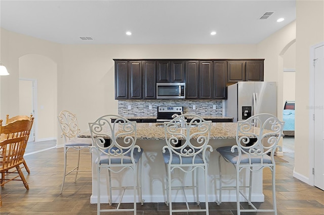 kitchen with light stone countertops, stainless steel appliances, tasteful backsplash, an island with sink, and a kitchen bar