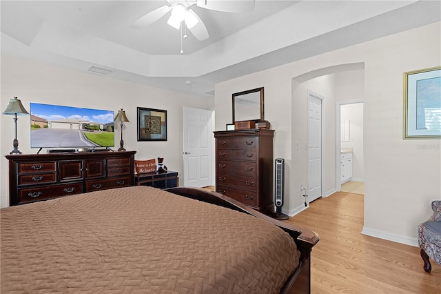 bedroom with light wood-type flooring, connected bathroom, a tray ceiling, and ceiling fan