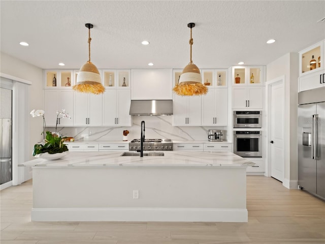 kitchen with hanging light fixtures, wall chimney exhaust hood, appliances with stainless steel finishes, and a kitchen island with sink