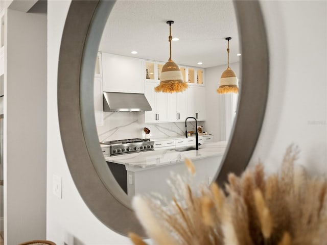 kitchen with decorative backsplash, wall chimney range hood, hanging light fixtures, stainless steel stove, and white cabinets