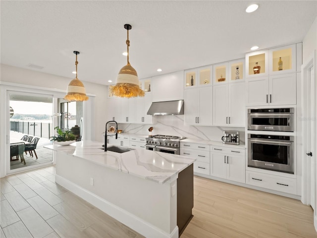 kitchen featuring exhaust hood, a center island with sink, light stone countertops, pendant lighting, and appliances with stainless steel finishes