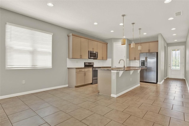 kitchen featuring light brown cabinetry, appliances with stainless steel finishes, an island with sink, decorative light fixtures, and a breakfast bar