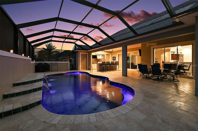 pool at dusk featuring a patio, a lanai, exterior bar, ceiling fan, and pool water feature