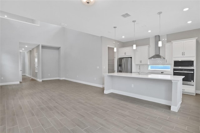 kitchen with appliances with stainless steel finishes, wall chimney exhaust hood, a center island with sink, light hardwood / wood-style floors, and white cabinetry