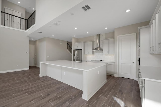 kitchen with stainless steel refrigerator with ice dispenser, wall chimney exhaust hood, white cabinets, a kitchen island with sink, and decorative backsplash