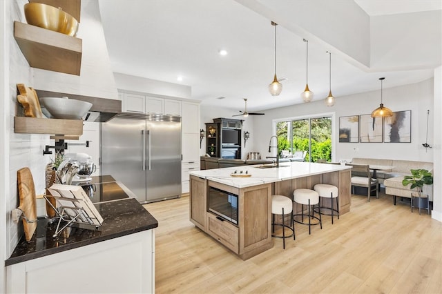 kitchen with built in appliances, an island with sink, dark stone countertops, light wood-type flooring, and white cabinetry