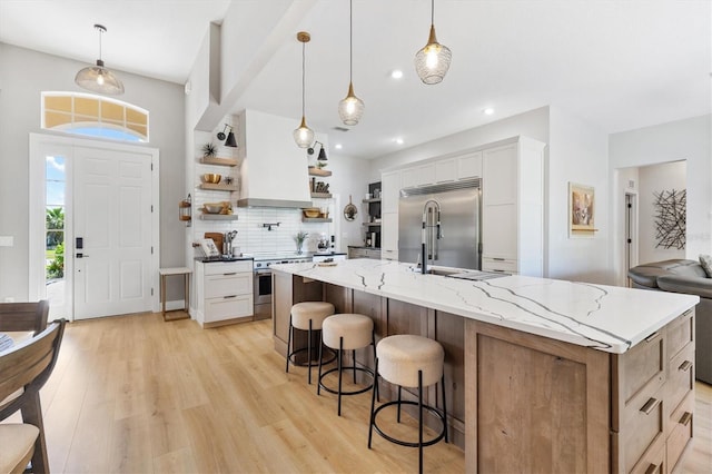 kitchen with white cabinets, hanging light fixtures, a large island with sink, appliances with stainless steel finishes, and light hardwood / wood-style flooring