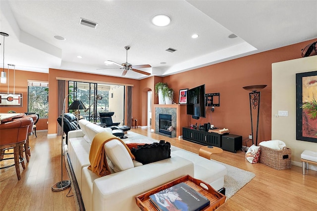 living room with light hardwood / wood-style flooring, a tray ceiling, a tile fireplace, a textured ceiling, and ceiling fan