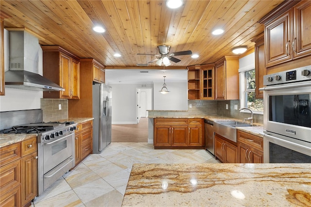 kitchen featuring wall chimney range hood, stainless steel appliances, sink, light stone countertops, and decorative light fixtures