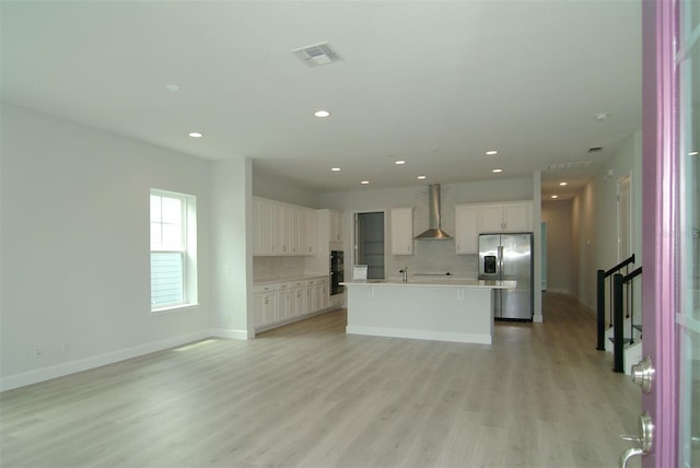 kitchen featuring white cabinets, wall chimney exhaust hood, stainless steel refrigerator with ice dispenser, and a kitchen island with sink