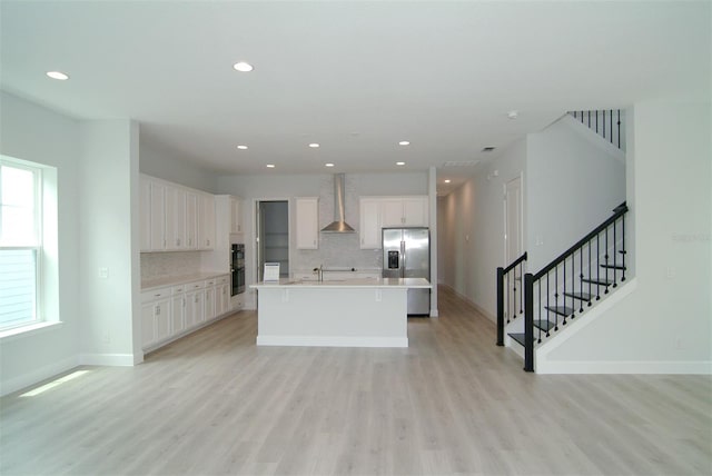 kitchen with wall chimney range hood, an island with sink, white cabinetry, light wood-type flooring, and stainless steel refrigerator with ice dispenser