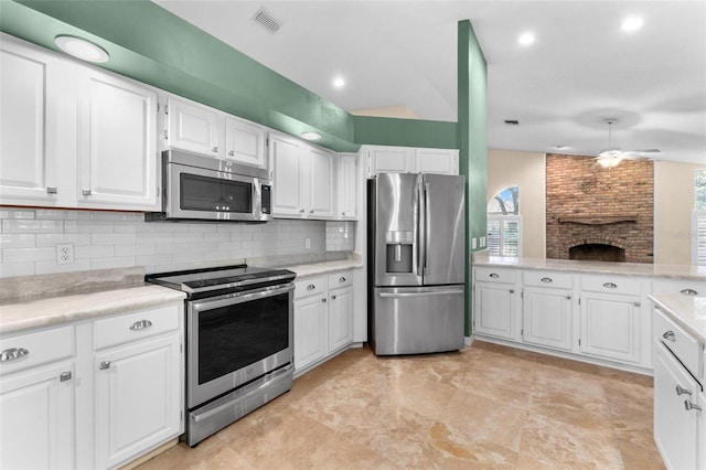 kitchen with decorative backsplash, stainless steel appliances, a brick fireplace, white cabinetry, and ceiling fan