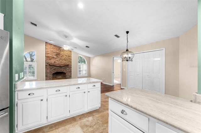 kitchen with vaulted ceiling, a brick fireplace, pendant lighting, white cabinets, and ceiling fan with notable chandelier