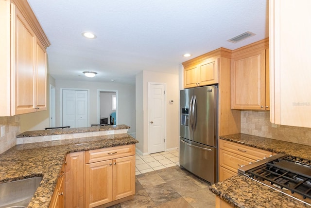 kitchen with stainless steel appliances, kitchen peninsula, dark stone counters, light brown cabinetry, and backsplash