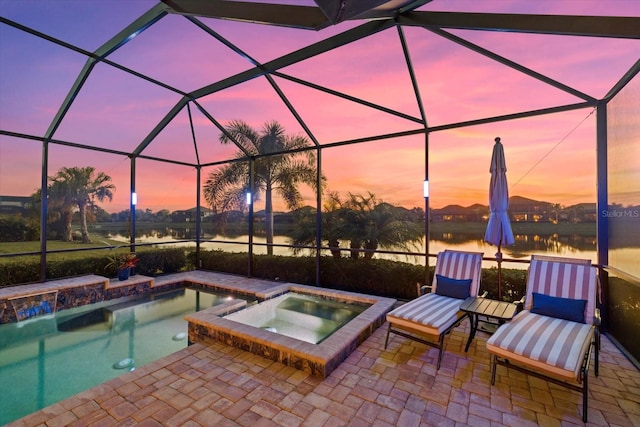 pool at dusk featuring a water view, a patio area, glass enclosure, and an in ground hot tub