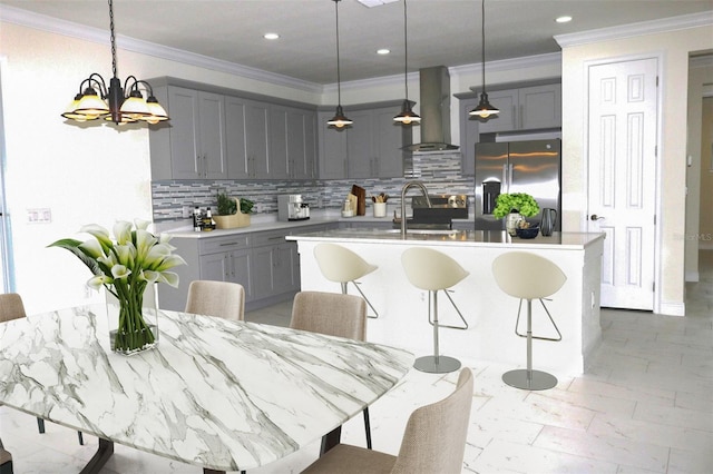 kitchen featuring wall chimney exhaust hood, stainless steel fridge with ice dispenser, hanging light fixtures, a center island with sink, and decorative backsplash