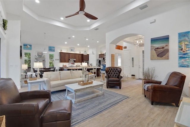 living room featuring ceiling fan with notable chandelier, a raised ceiling, plenty of natural light, and light hardwood / wood-style flooring