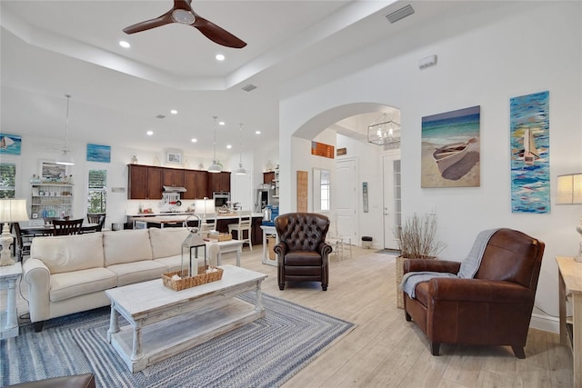 living room featuring light wood-type flooring, ceiling fan with notable chandelier, and a tray ceiling