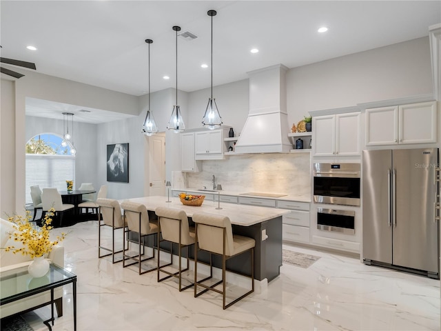 kitchen featuring white cabinets, hanging light fixtures, an island with sink, custom range hood, and stainless steel appliances