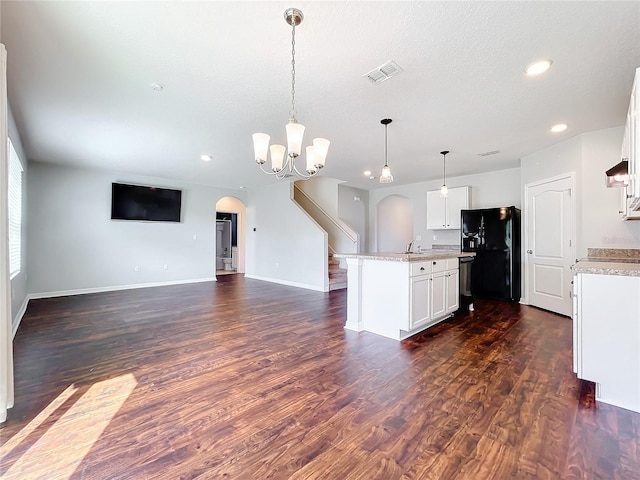 kitchen featuring dark wood-type flooring, white cabinets, black fridge, an island with sink, and decorative light fixtures